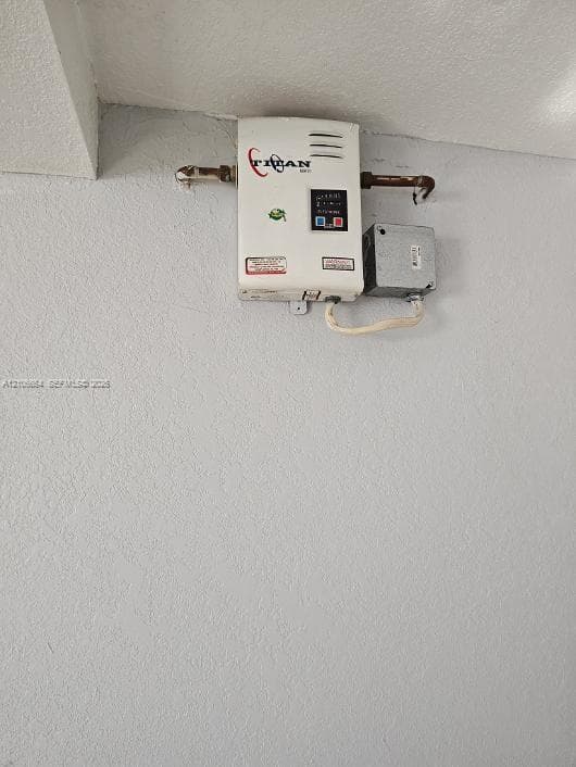 Wall-mounted tankless water heating unit featuring a digital temperature control panel, metal pipe connections, and a dedicated electrical junction box