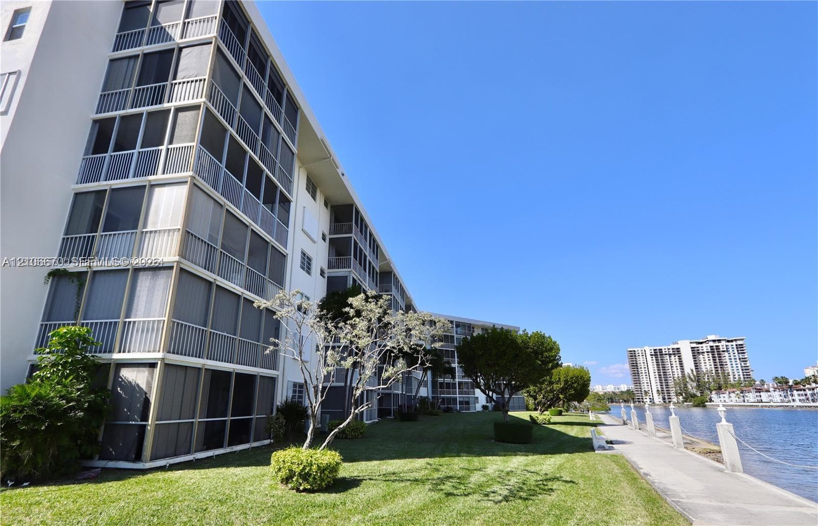 Multi-story building exterior featuring screened-in balconies, concrete walkway, manicured lawn, and water frontage