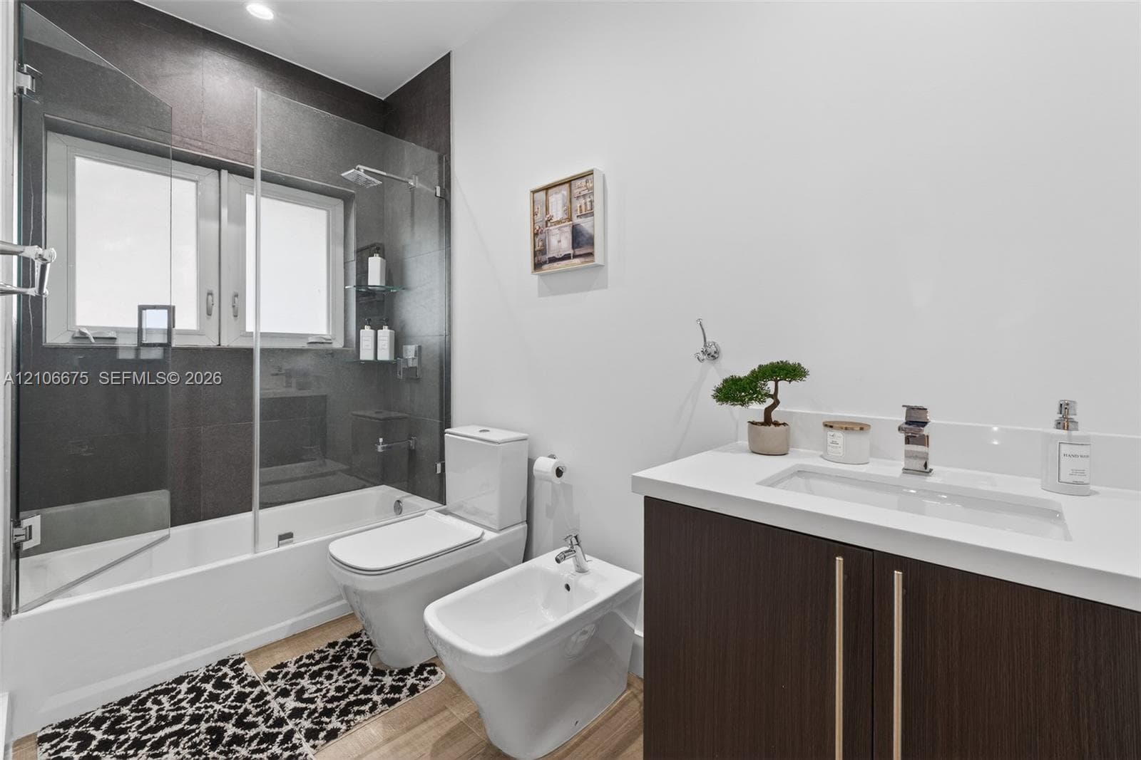 Bathroom featuring a dark wood-finish vanity with a white countertop and rectangular sink, frameless glass shower enclosure, and dark tile wall surround