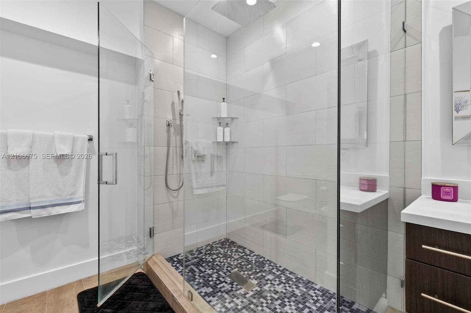 Walk-in shower featuring a glass enclosure, rainfall showerhead, and mosaic tile flooring, complemented by a dark wood-finish vanity with a white countertop