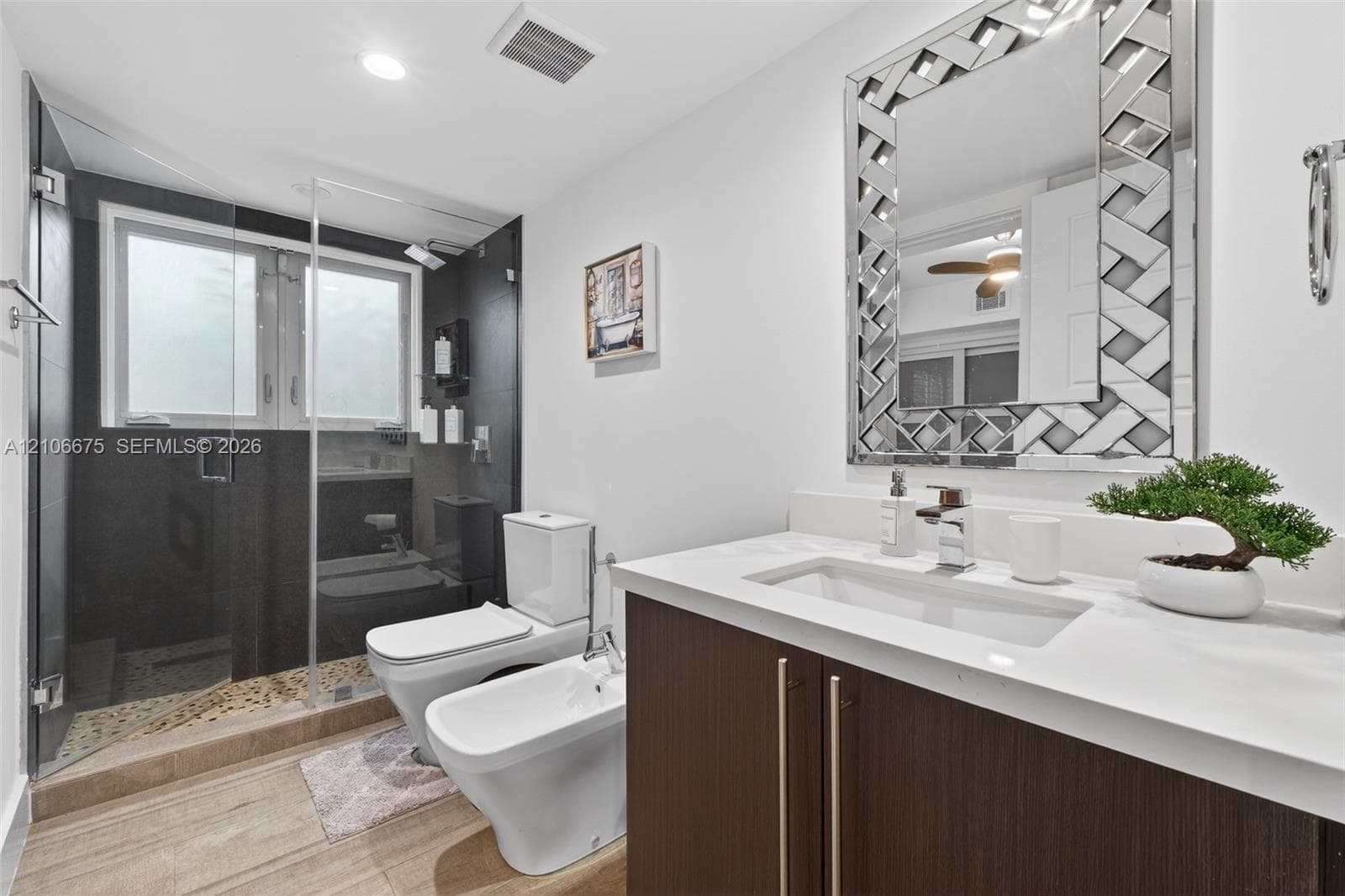 Bathroom featuring a vanity with dark wood-finish cabinetry, a white countertop with an integrated rectangular sink, a glass-enclosed shower with dark tile surround, a wall-mounted mirror with a decorative frame, and stone-finish flooring