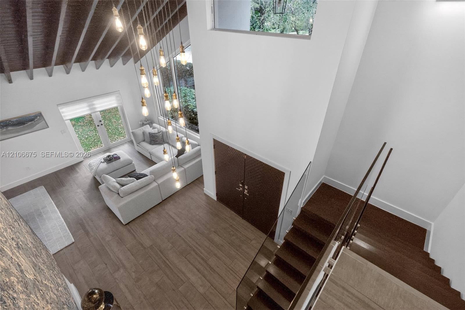Double-height ceiling with exposed wooden rafters, expansive multi-pane windows, wood-finish flooring, staircase featuring a glass panel railing, and a multi-pendant light fixture