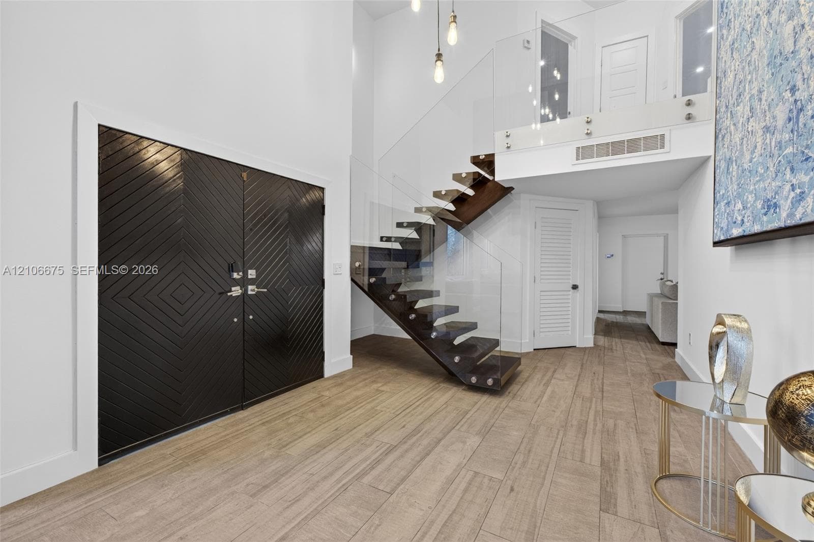 Double-height entryway featuring wood-finish tile flooring, a geometric patterned double door, and a floating staircase with glass panel railings and pendant lighting