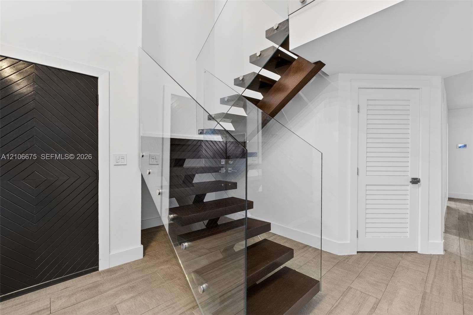 Floating staircase with dark wood-finish treads and frameless glass railing, accented by a geometric patterned door and louvered door, set over wood-finish tile flooring