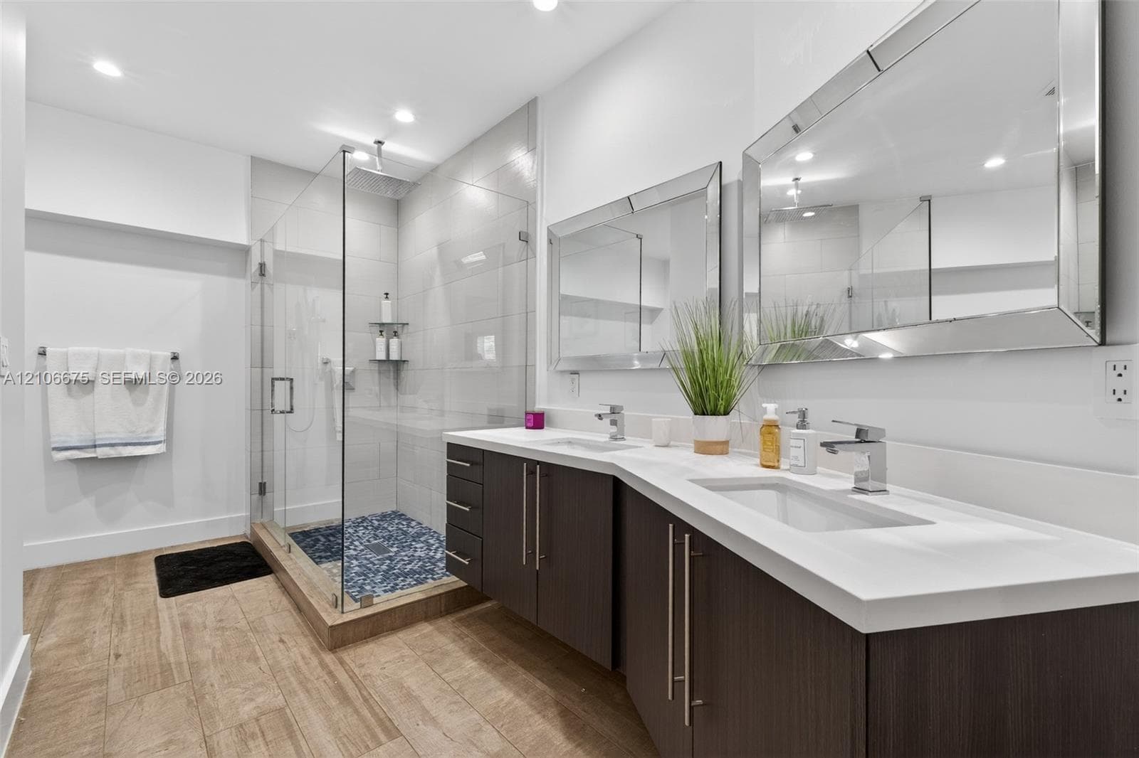 Dual-sink vanity with a white countertop and dark-finish cabinetry, frameless glass shower enclosure with stone tile floor, and large rectangular wall-mounted mirrors