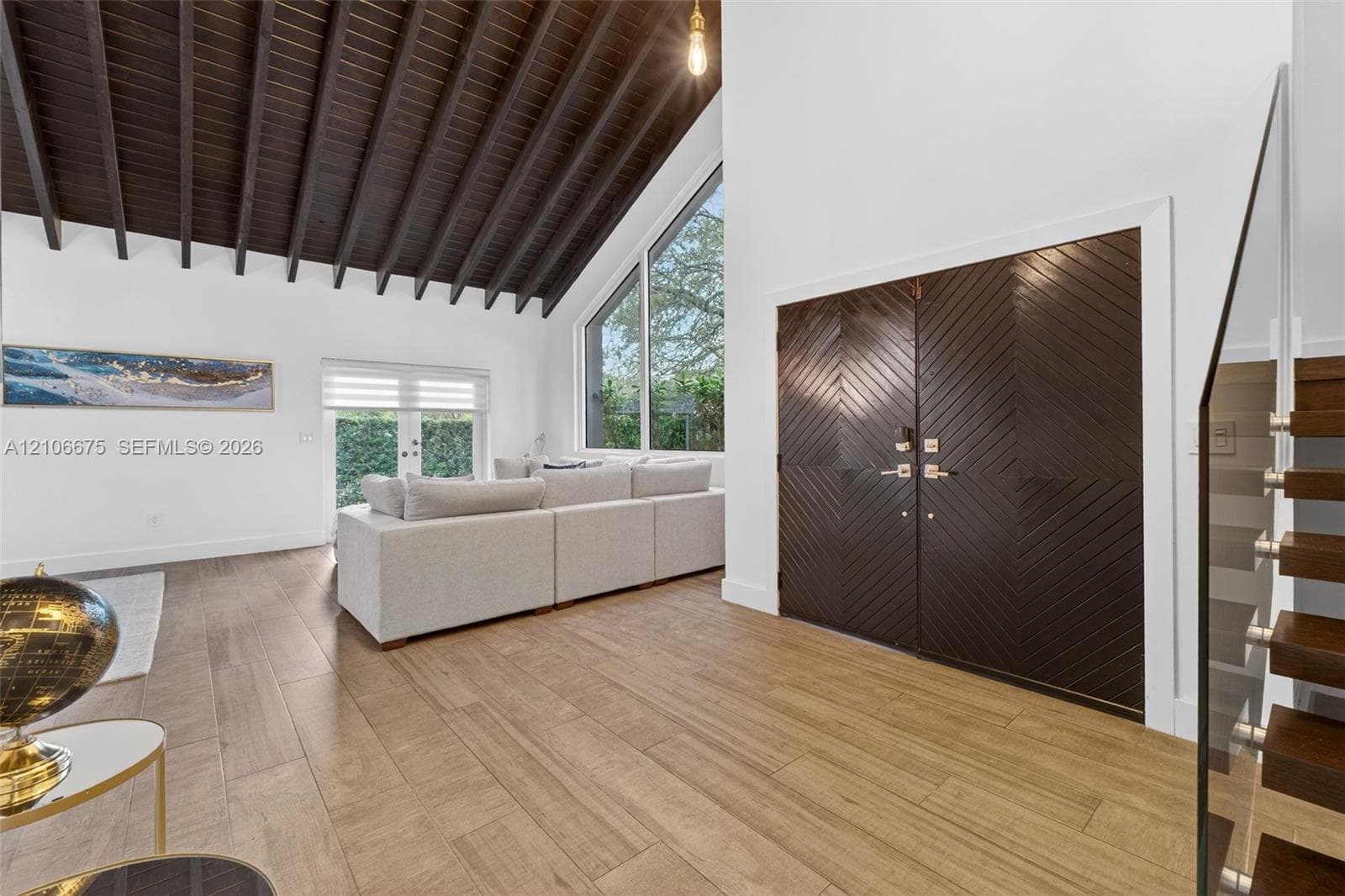 Vaulted ceiling with exposed dark wood-finish beams, expansive floor-to-ceiling geometric windows, wood-finish flooring, double doors featuring chevron-patterned woodwork, and a glass-paneled staircase railing