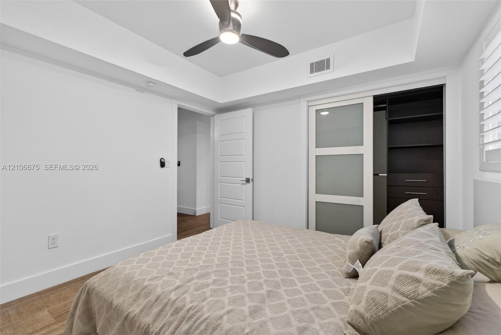 Tray ceiling with integrated ceiling fan, frosted-glass sliding closet doors, built-in closet shelving, wood-finish flooring, and shutter window coverings