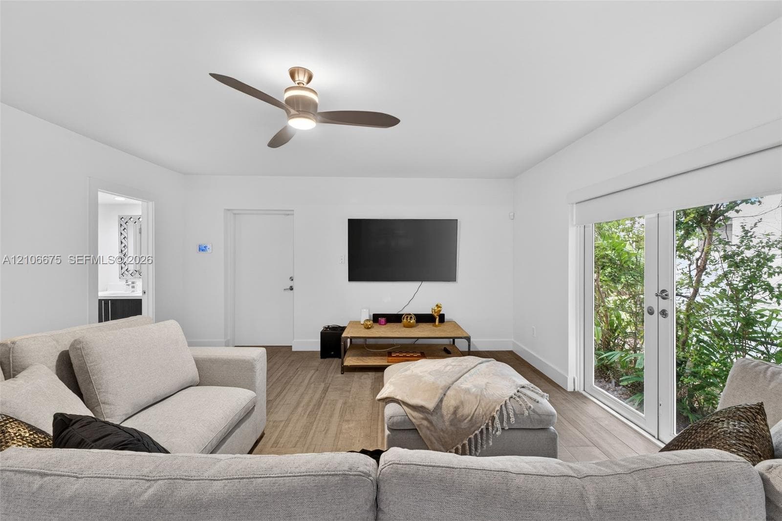 Neutral-toned interior space featuring wood-finish flooring, a modern multi-blade ceiling fan with integrated lighting, and full-height glass French doors