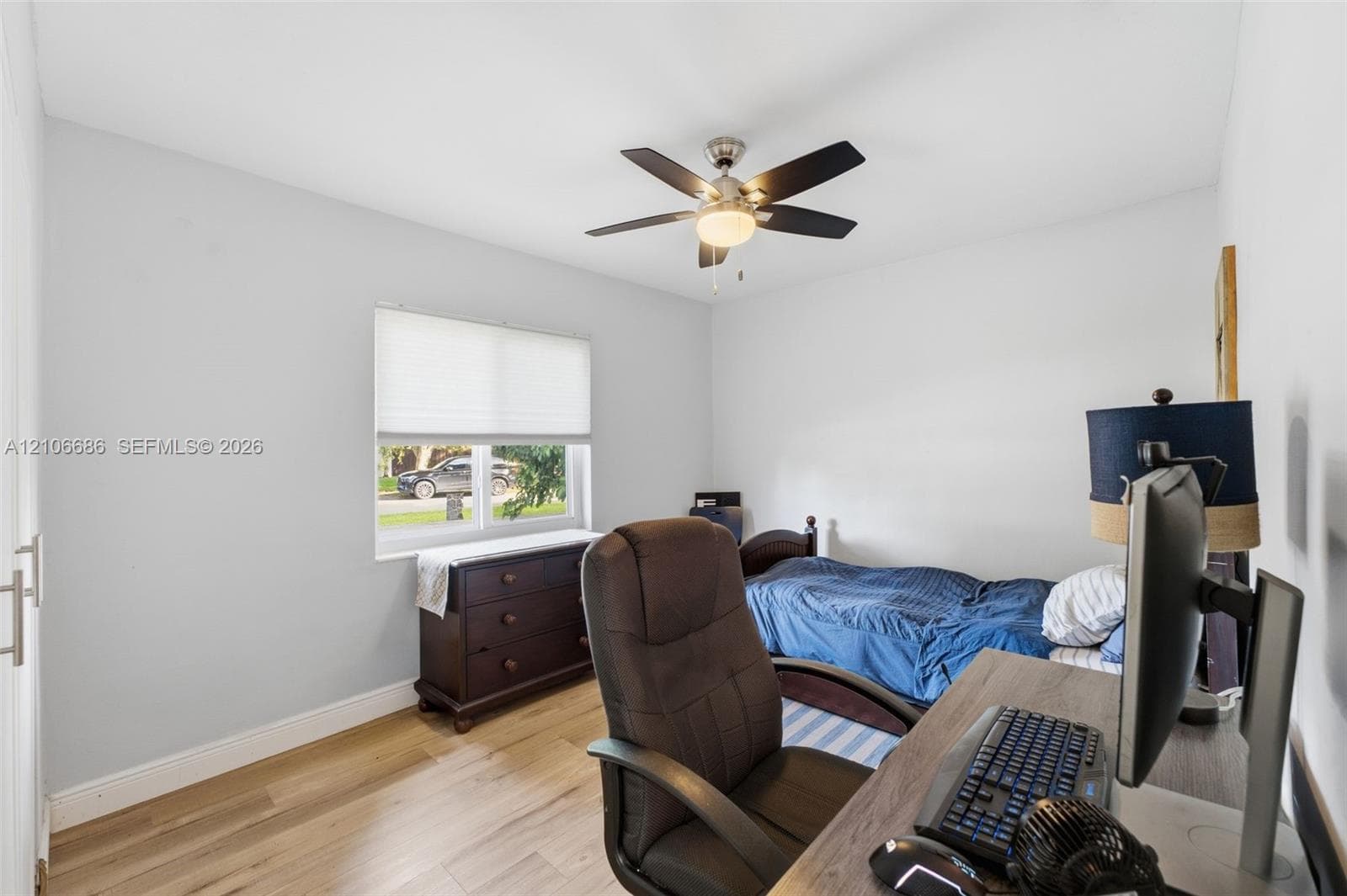 Room featuring wood-finish flooring, a multi-blade ceiling fan with integrated lighting, and a window with roller shade