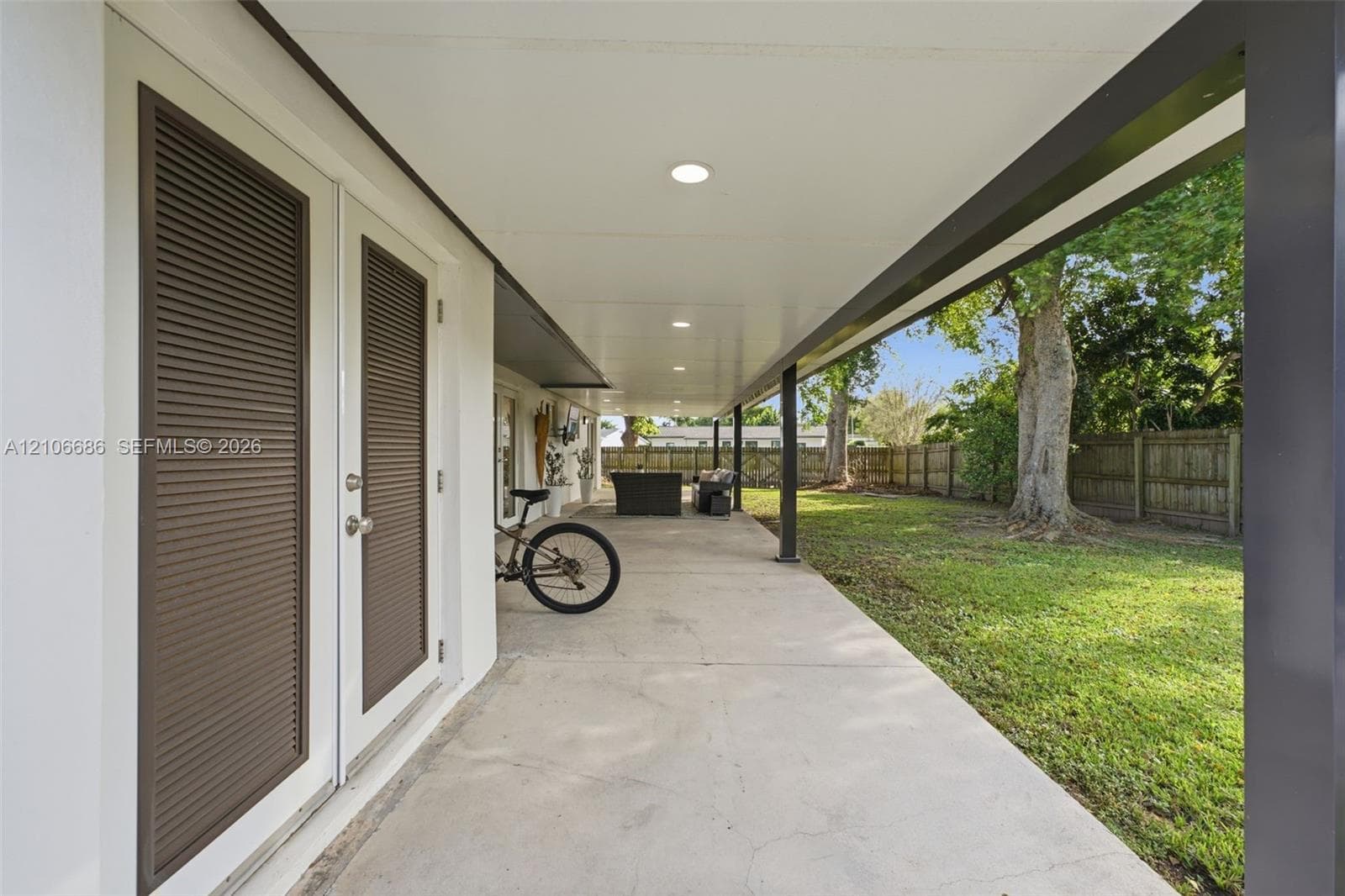 Covered concrete patio featuring recessed lighting, double doors with louvered panels, and metal support posts overlooking a fenced backyard with mature trees