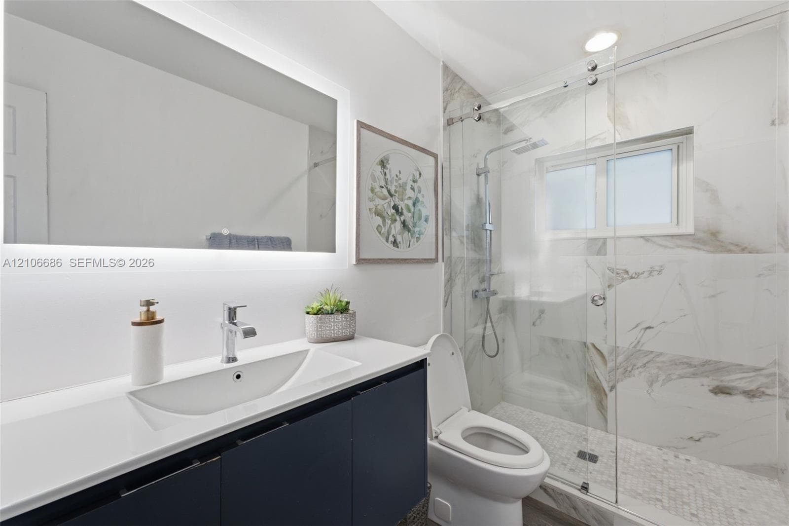 Bathroom featuring a dark vanity with a white countertop, integrated sink, illuminated wall mirror, and walk-in shower with stone-finish tiling and a glass enclosure