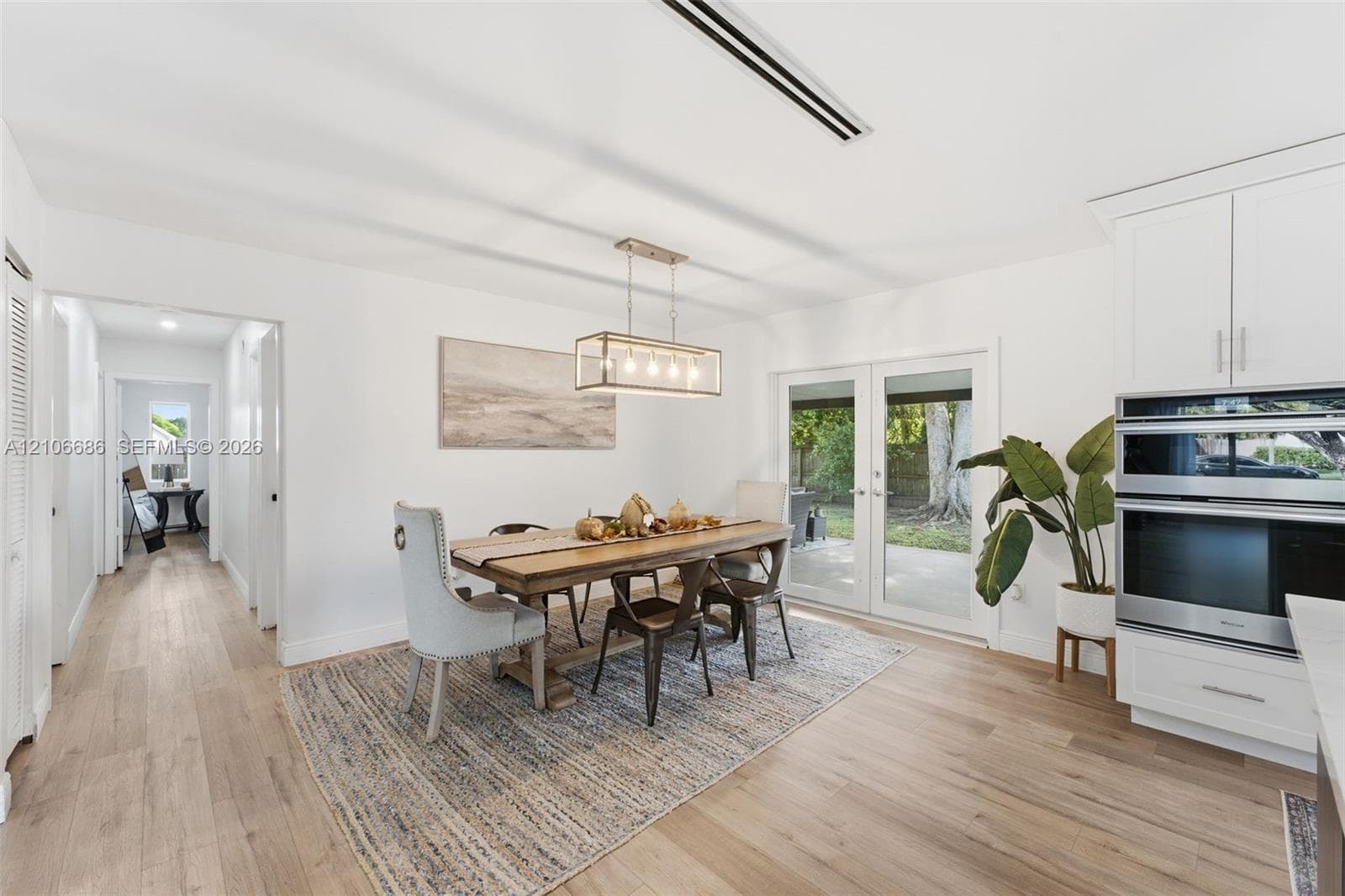 Dining area featuring light-toned wood-finish flooring, white shaker-style cabinetry, double wall-mounted ovens, a rectangular pendant light fixture, and glass-paned double doors