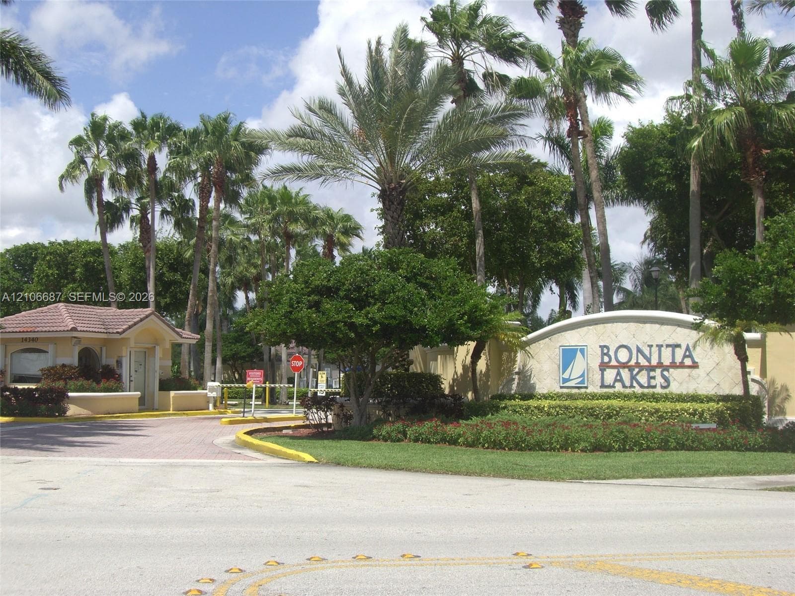 Gated community entrance featuring a guardhouse with a tiled roof, stone-clad monument signage, and mature palm tree landscaping