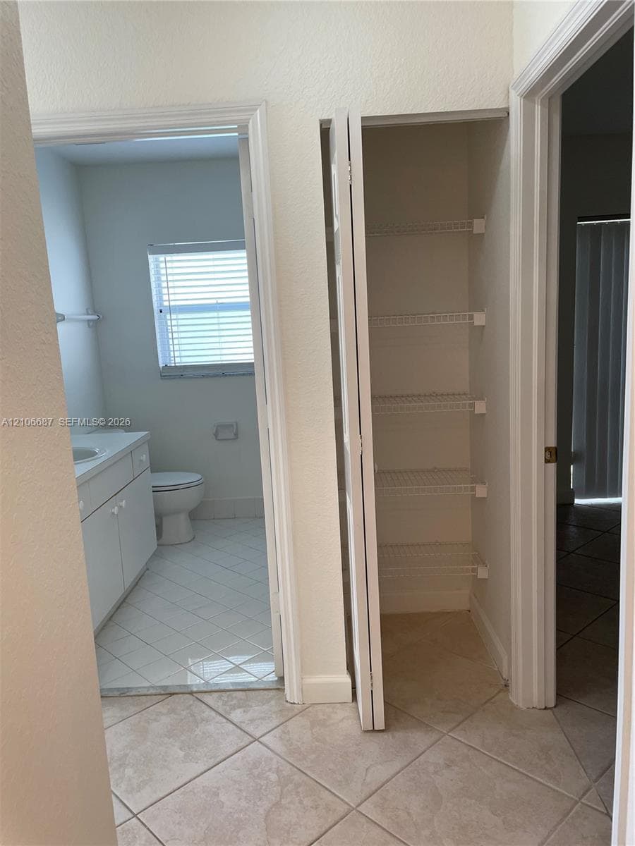 Interior hall area featuring ceramic tile flooring, a floor-to-ceiling closet with ventilated wire shelving, and a bathroom entry containing a white vanity with an integrated sink and a porcelain toilet