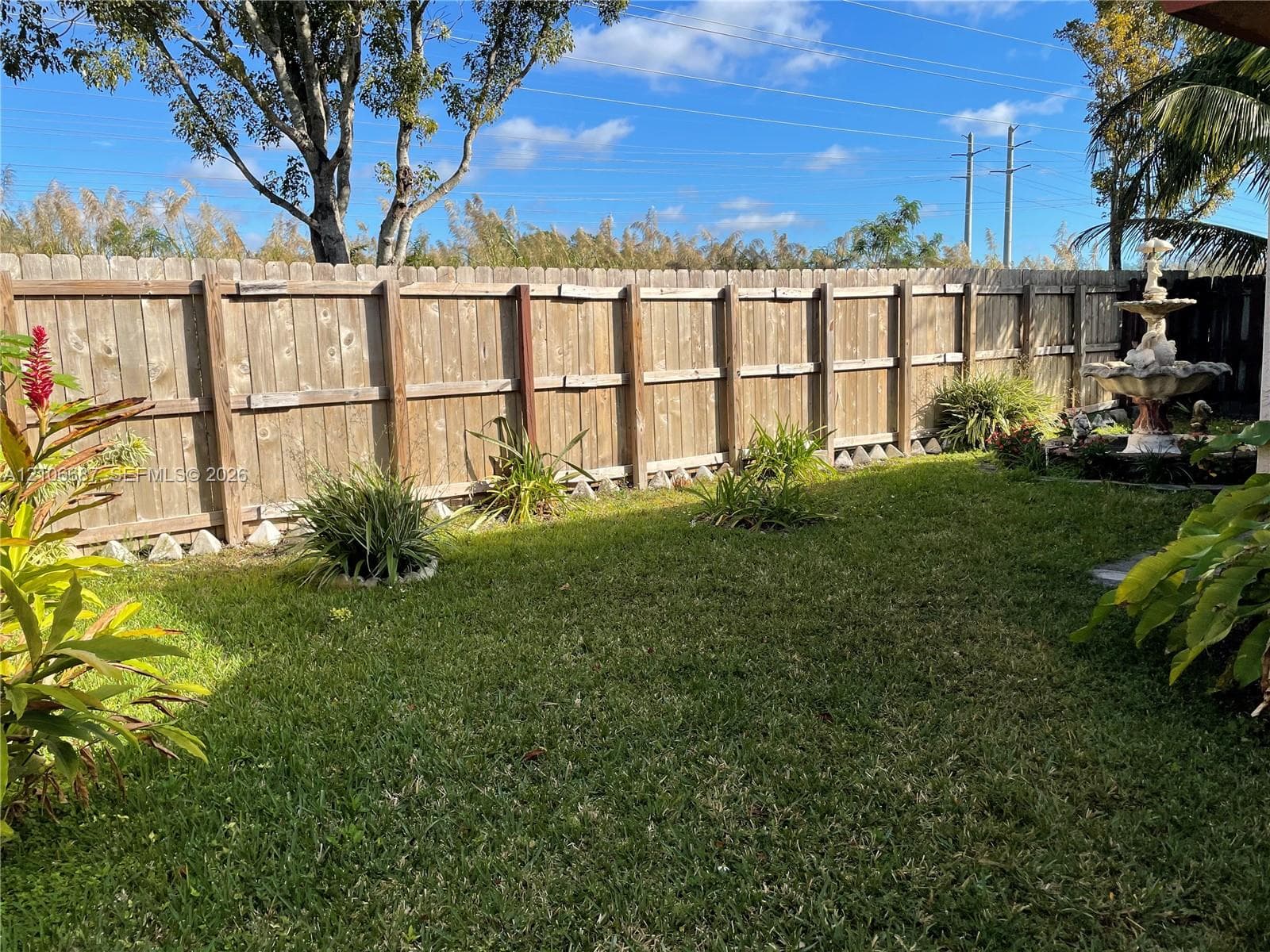 Private backyard featuring a wood-panel fence, manicured lawn, established landscaping, and a stone-finish tiered water fountain
