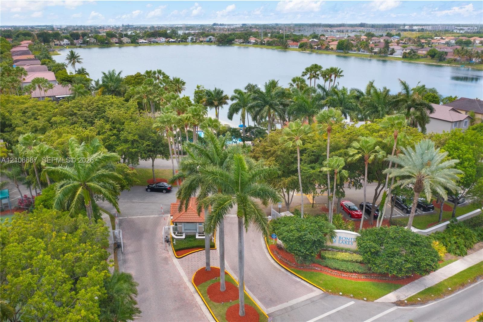 Gated community entrance featuring a guardhouse, mature palm tree landscaping, paved access roads, and direct lake frontage