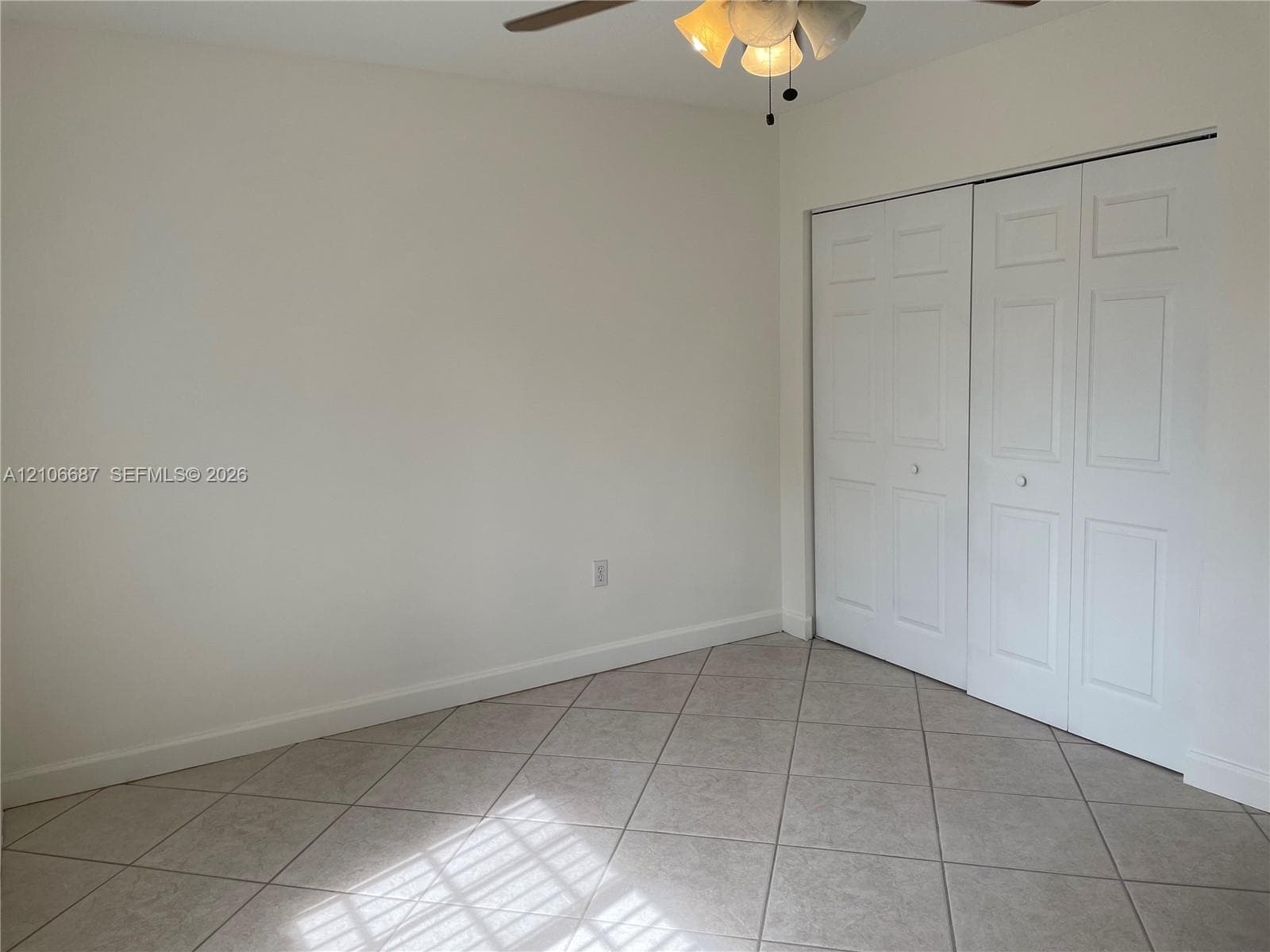 Neutral-toned tile flooring set on a diagonal pattern, white paneled bi-fold closet doors, and a ceiling-mounted light fixture with a fan