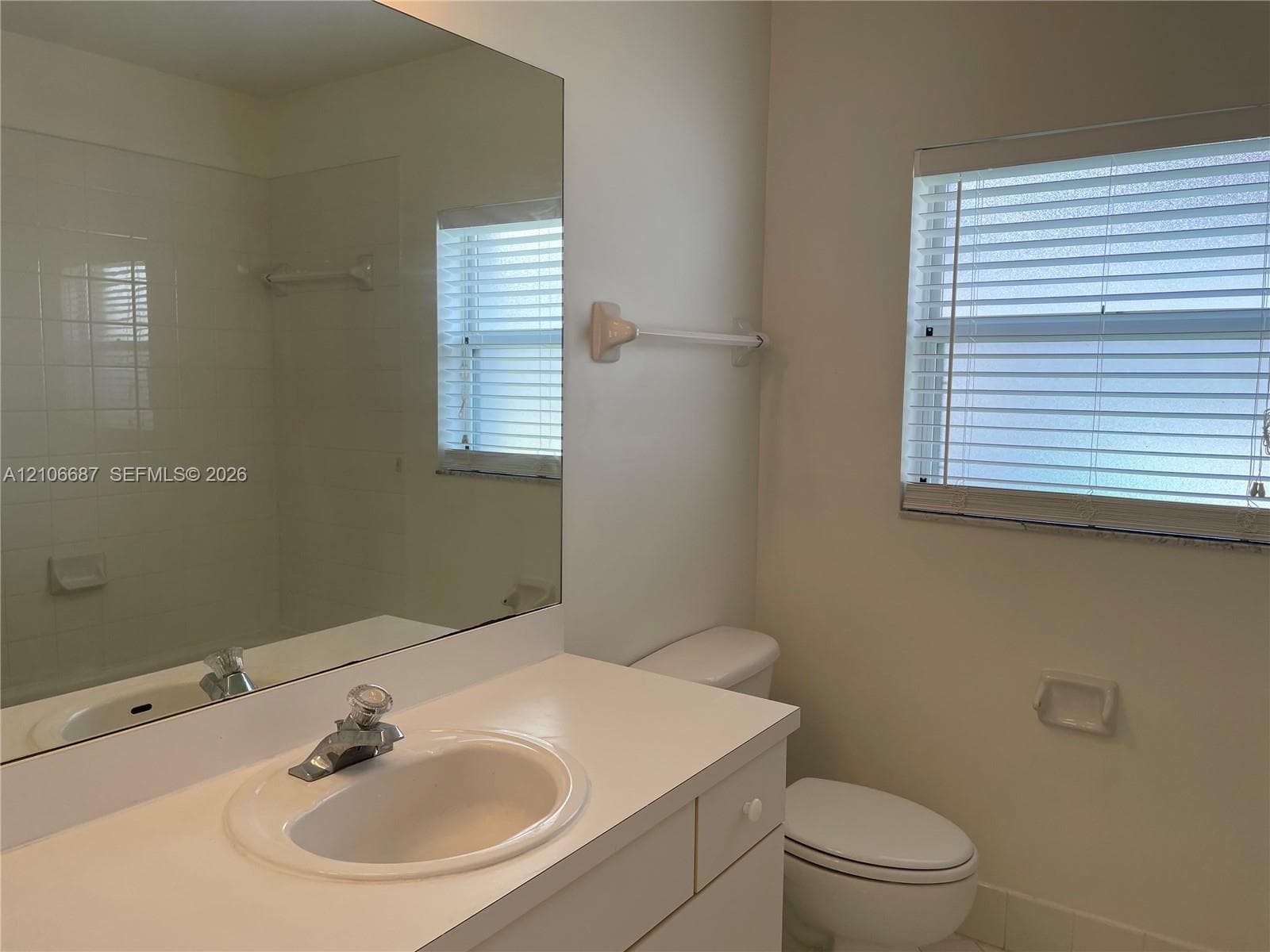 Bathroom featuring a white vanity with an integrated sink, a large wall-mounted mirror, a tiled shower surround, and a window with horizontal blinds