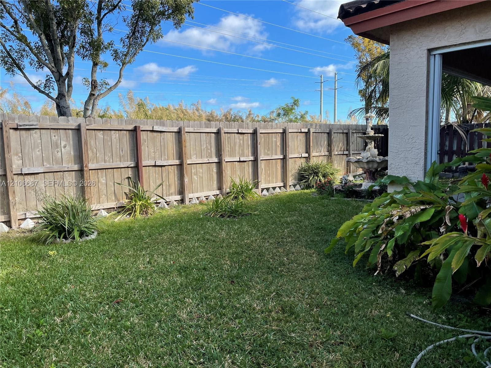 Private backyard featuring a fenced perimeter, grass lawn, mature trees, and tiered stone fountain