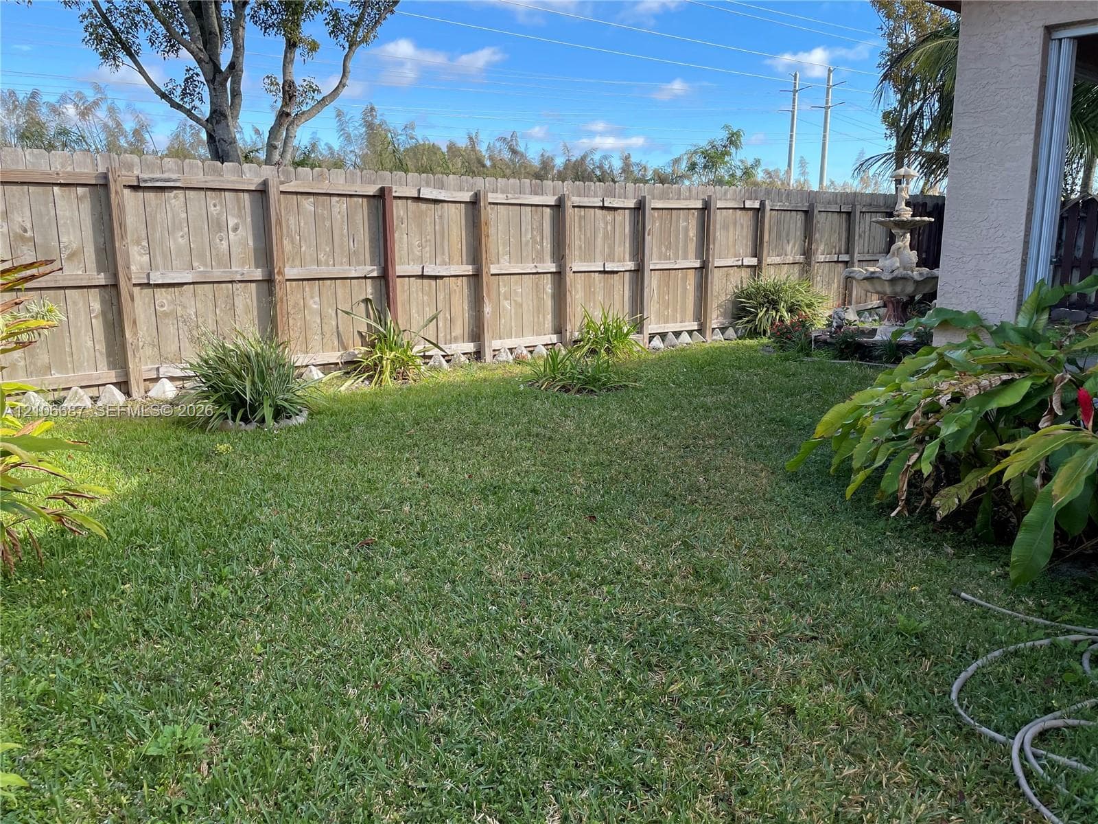 Rear yard featuring a grass lawn, wood-panel perimeter fencing, decorative rock border, mature greenery, and a tiered stone fountain