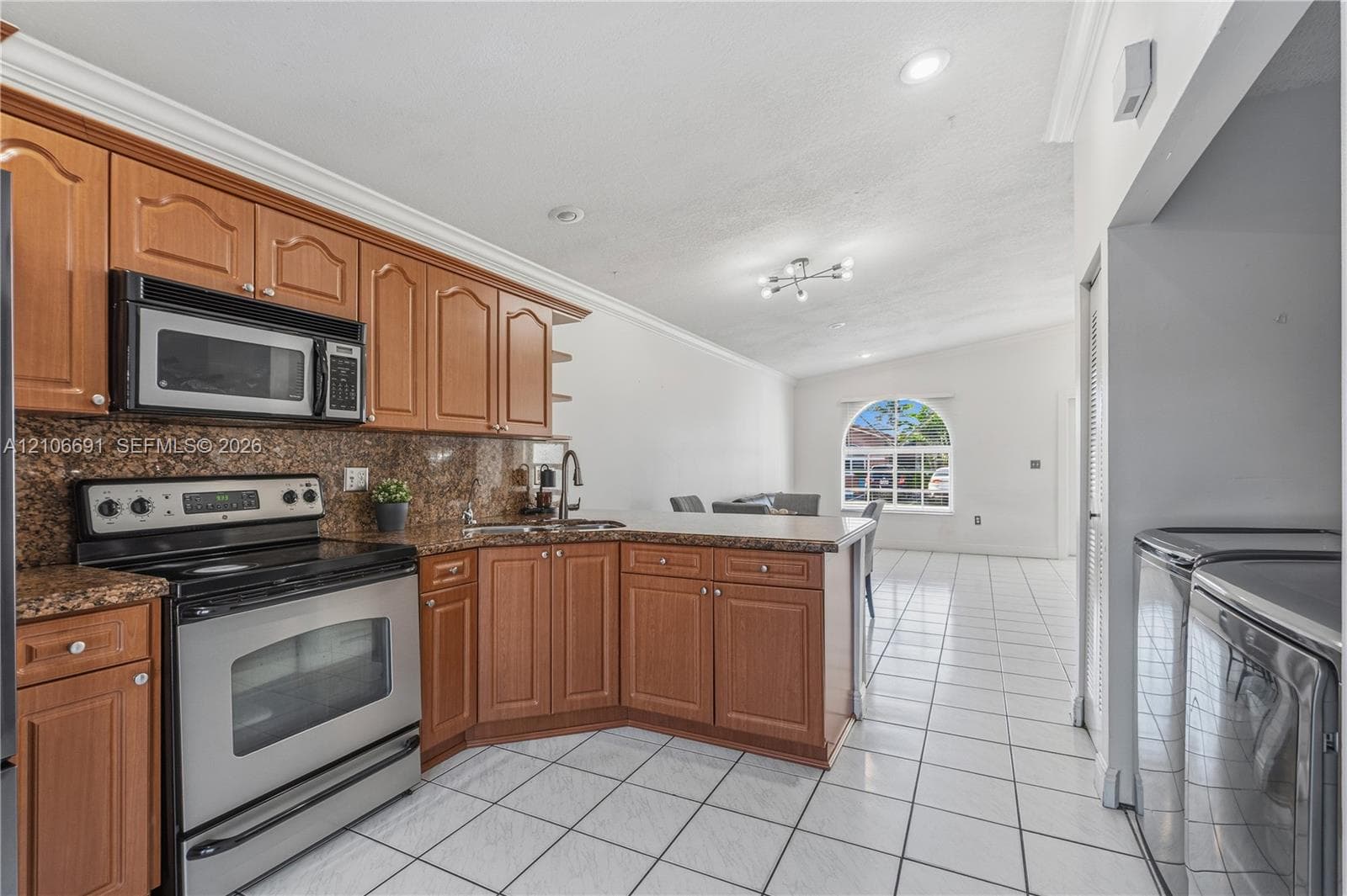 Kitchen featuring medium-tone wood cabinetry, stone countertops, stainless steel appliances, tile flooring, and a multi-light ceiling fixture
