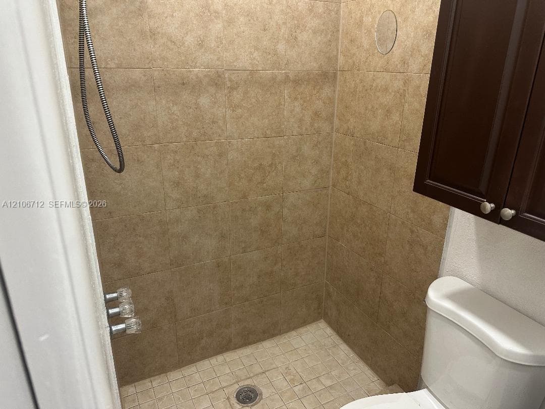 Walk-in shower featuring floor-to-ceiling tile surround, mosaic tile flooring, and metallic fixtures, accompanied by an elevated dark wood-finish cabinet and porcelain commode
