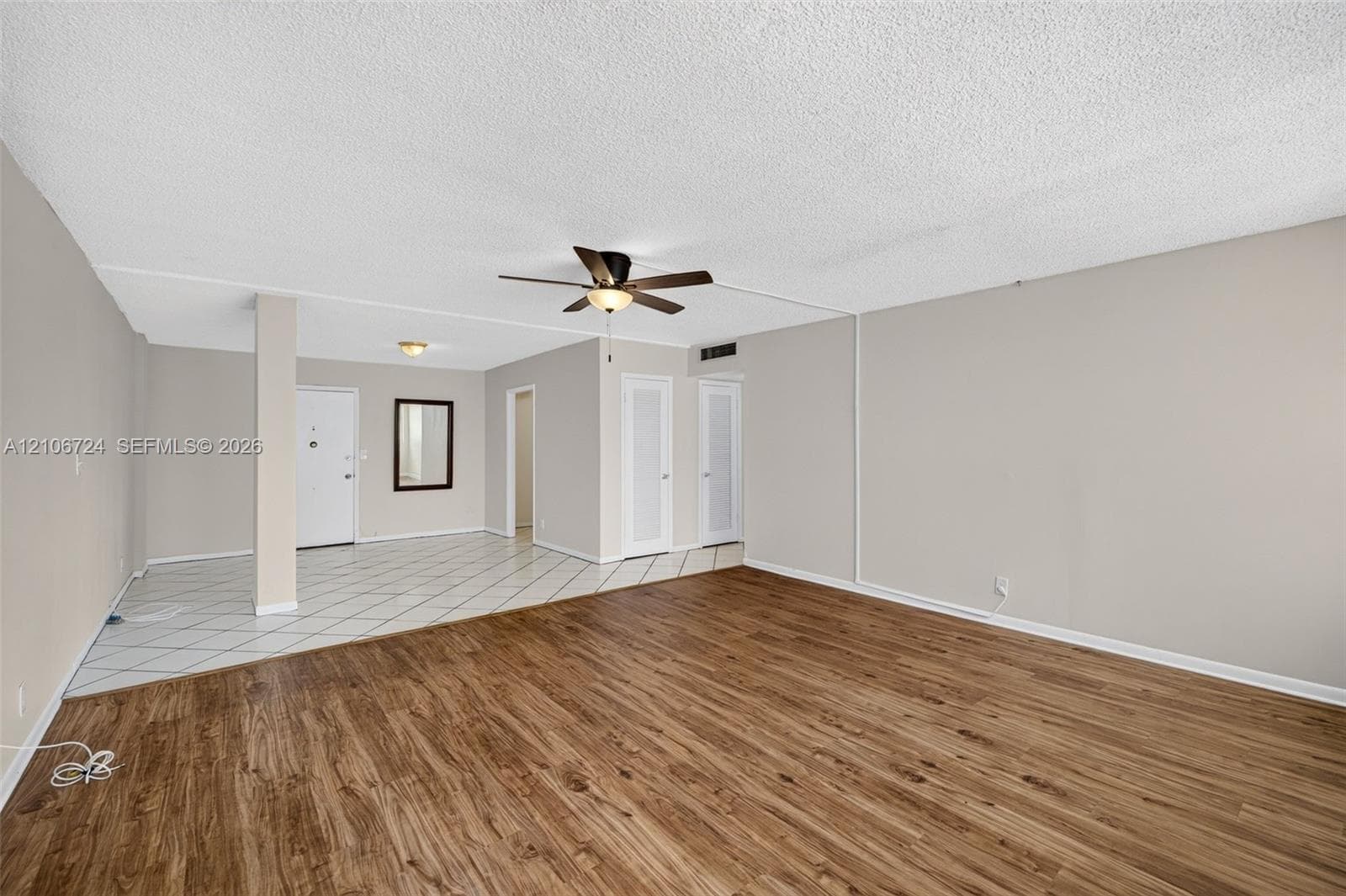 Open-concept interior featuring wood-finish flooring, ceramic tile flooring, a central structural support pillar, a ceiling fan with integrated lighting, and dual closet doors