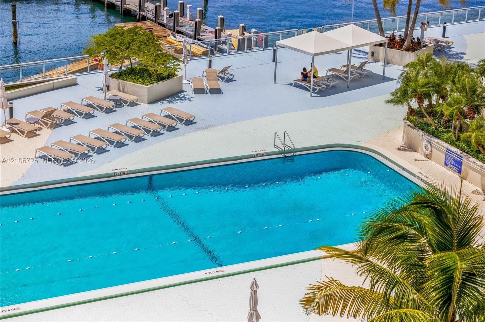 In-ground swimming pool with lane markers, paved deck, overhead poolside canopies, concrete planters with mature landscaping, and waterfront dock access