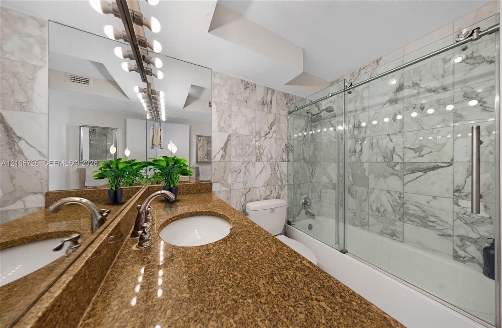 Bathroom featuring an expansive vanity with a dual-toned stone countertop, dual undermount sinks, a full-width mirror with integrated lighting, and a bathtub with a stone-tile surround and a glass sliding enclosure