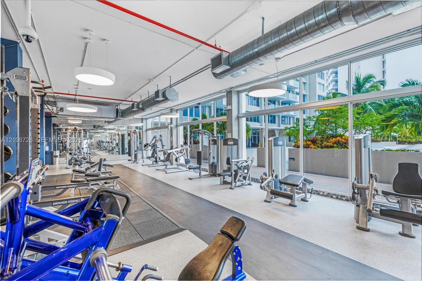 Fitness center featuring floor-to-ceiling windows, exposed industrial-style ductwork, recessed drum pendant lighting, and rubberized flooring