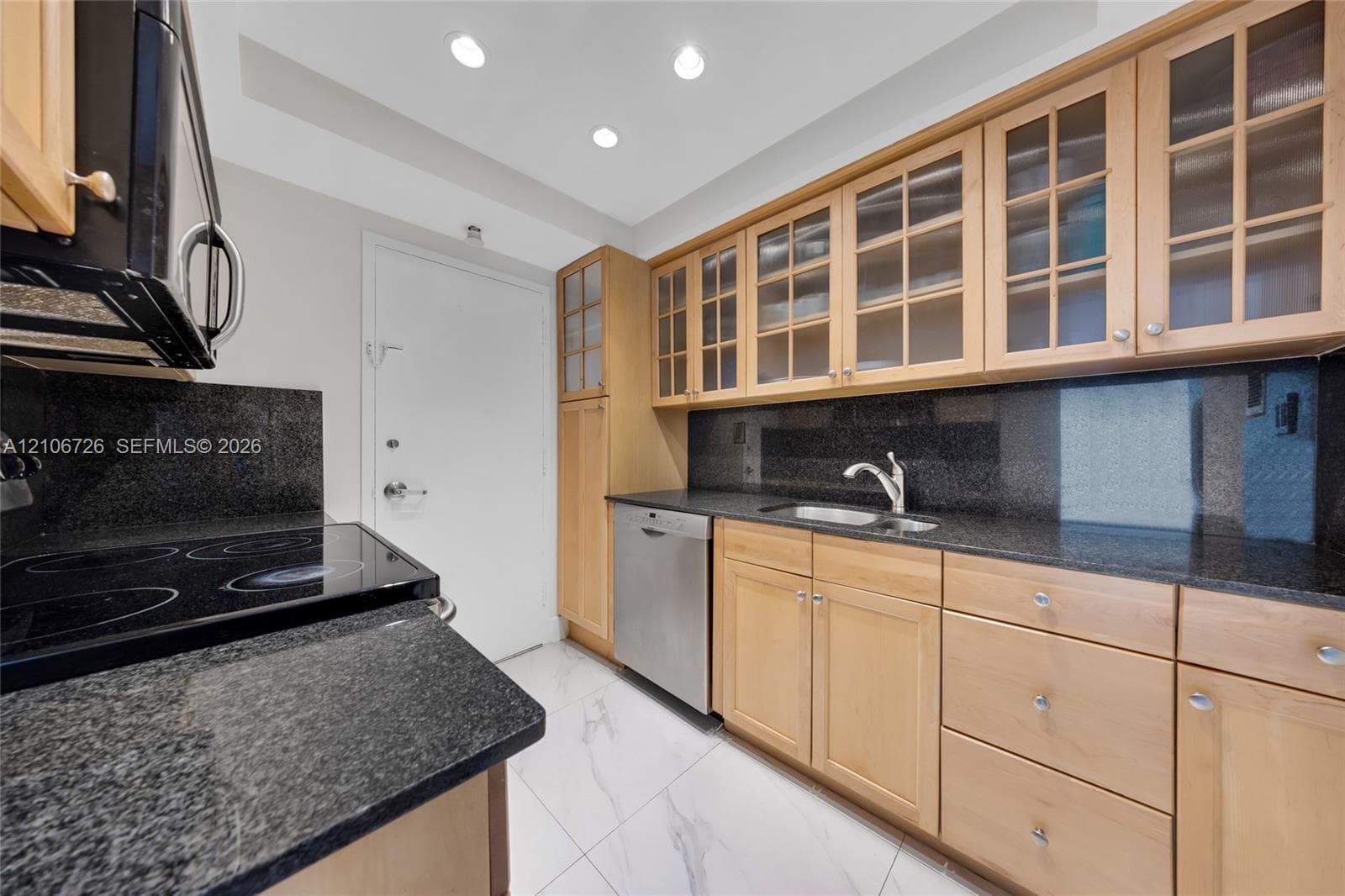 Kitchen featuring light wood cabinetry with glass-front upper doors, dark stone countertops, stainless steel dishwasher, integrated sink with gooseneck faucet, and white stone-finish flooring