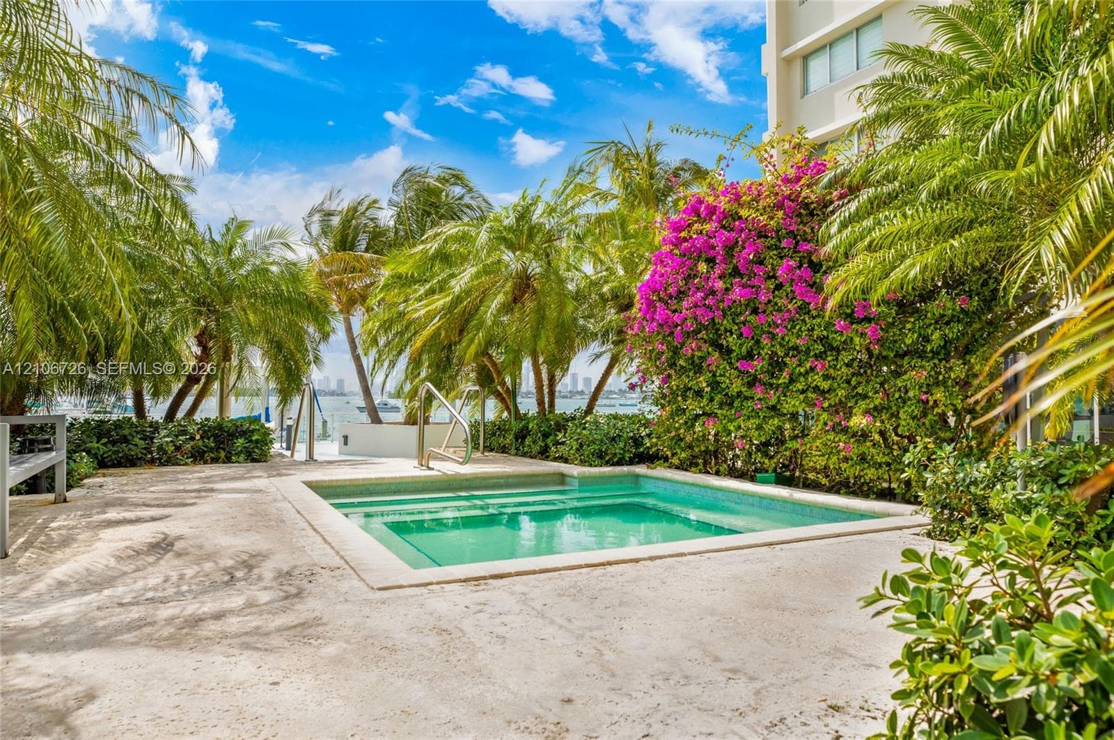 In-ground swimming pool featuring a concrete deck, built-in steps, and metal handrail, surrounded by mature palm trees and flowering shrubbery