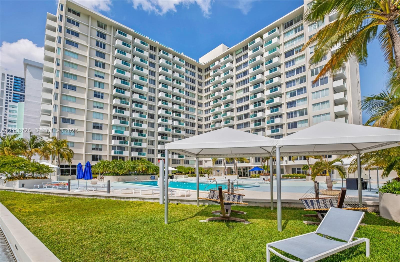 Mid-rise residential building featuring private balconies, expansive swimming pool, landscaped lawn, and white structural canopy pavilions
