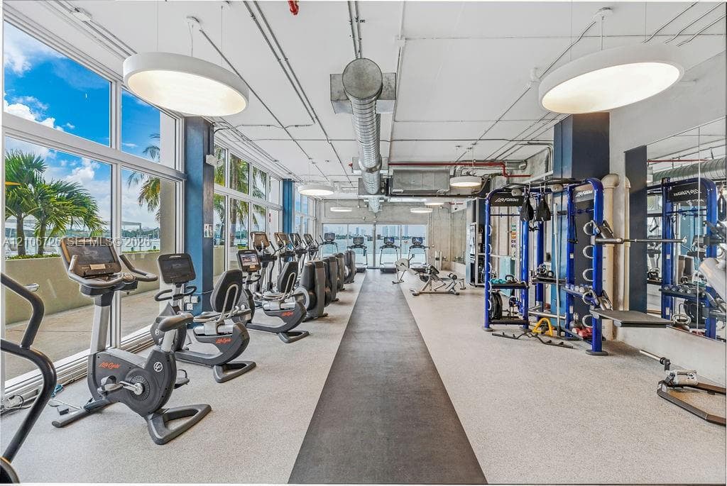 Fitness facility featuring floor-to-ceiling windows, exposed industrial-style ductwork, recessed circular lighting, rubberized flooring, and mirrored walls