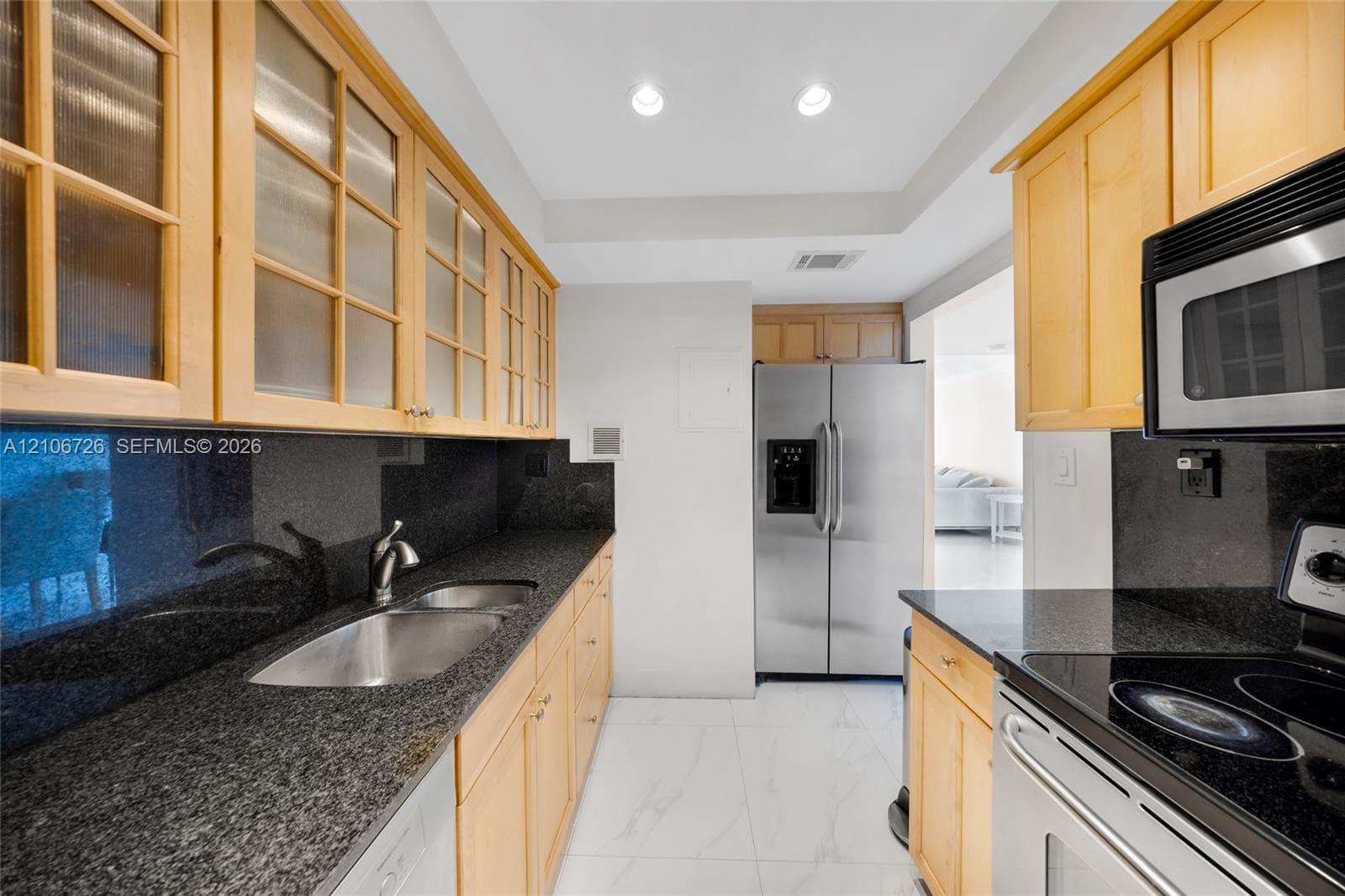 Kitchen featuring light-tone cabinetry with glass-paneled upper doors, dark stone countertops, stainless steel appliances, and large-format tile flooring