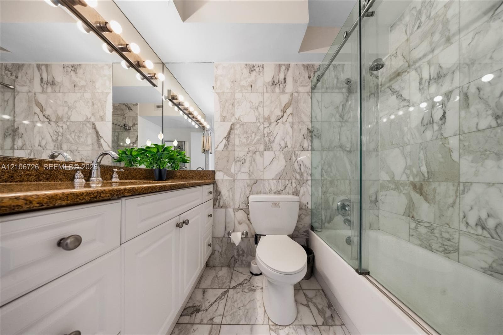 Full-width wall mirror with integrated multi-bulb lighting, white cabinetry with metallic hardware, stone-finish countertop, and full-height stone tile surround in the tub-shower combination with a sliding glass door