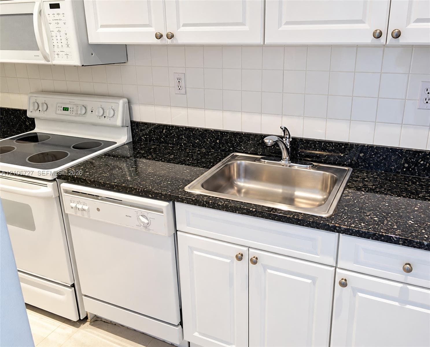 Kitchen featuring white cabinetry, dark speckled stone-surface countertops, a stainless steel undermount sink with a chrome faucet, a white square-tile backsplash, and a white built-in dishwasher