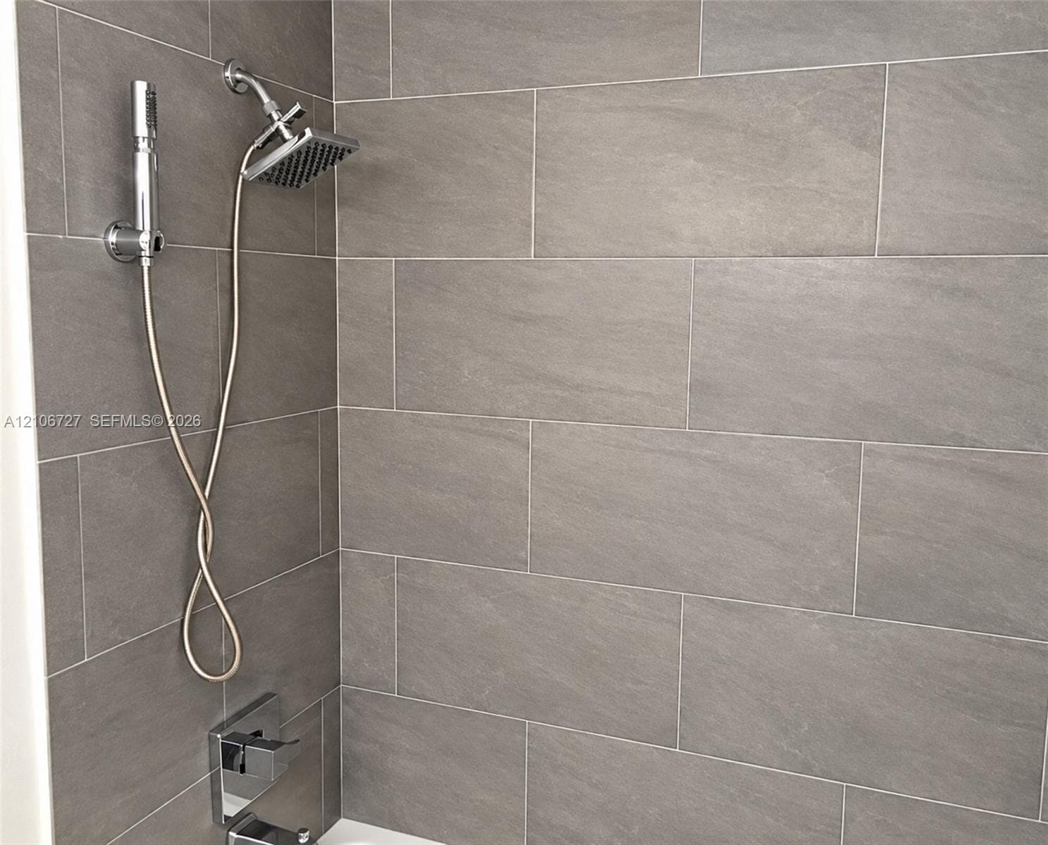 Shower area featuring large-format grey tile wall cladding, a wall-mounted rainfall shower head, a handheld shower wand attachment, and a polished chrome rectangular tub spout