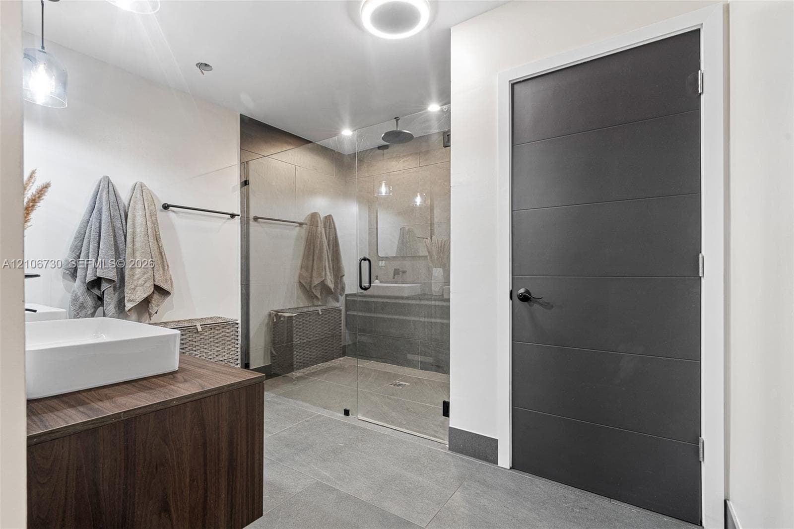 Bathroom featuring a wood-finish vanity with a white vessel sink, large-format tile flooring, a glass-enclosed walk-in shower, and a matte black hardware package