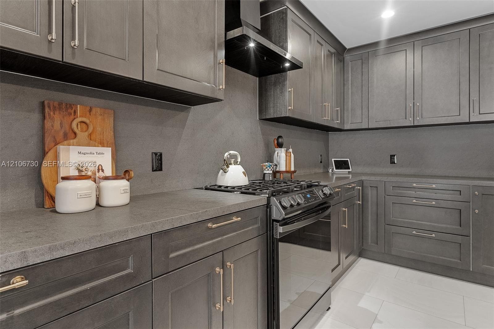 Kitchen featuring charcoal-toned cabinetry, solid-surface countertops with integrated backsplash, stainless steel gas range, black range hood, and large-format tile flooring