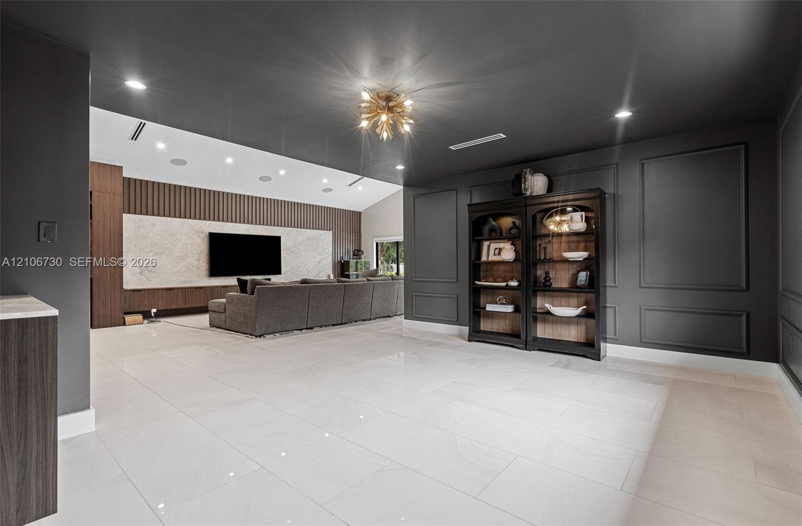 Large format tile flooring, charcoal-painted walls with decorative wainscoting, statement gold-tone ceiling fixture, recessed lighting, and a custom media wall featuring natural stone slab cladding and wood-panel detailing
