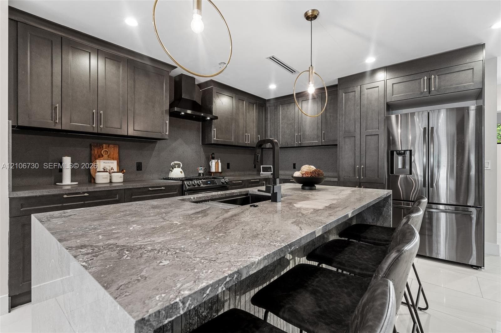 Kitchen featuring dark custom cabinetry, a stone-topped waterfall island, stainless steel appliances, a black range hood, and modern pendant lighting