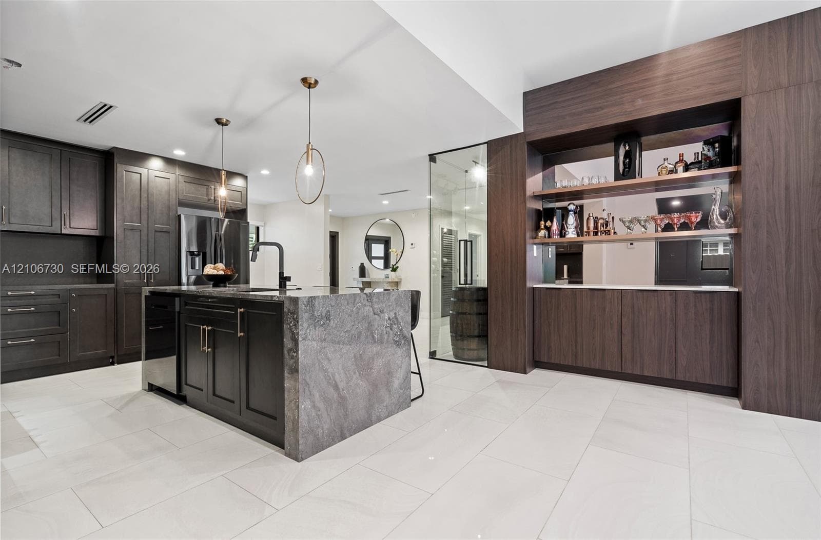 Kitchen featuring dark wood-finish cabinetry, a waterfall-edge stone island, built-in shelving with mirrored backsplash, and large-format tile flooring