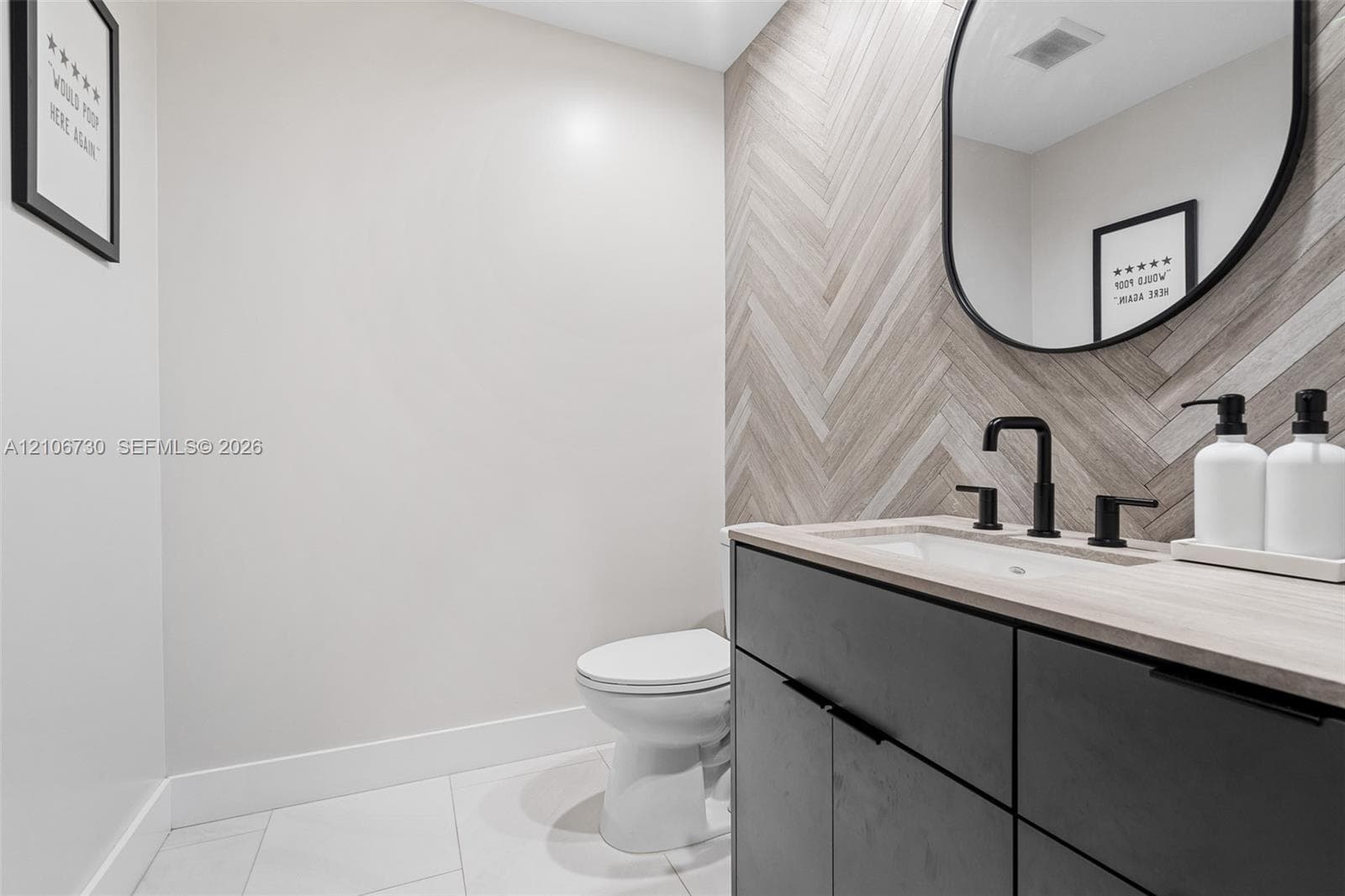 Herringbone pattern wall tile, charcoal-toned vanity cabinetry, light-toned countertop with integrated sink, matte black widespread faucet, and oval wall mirror with black framing