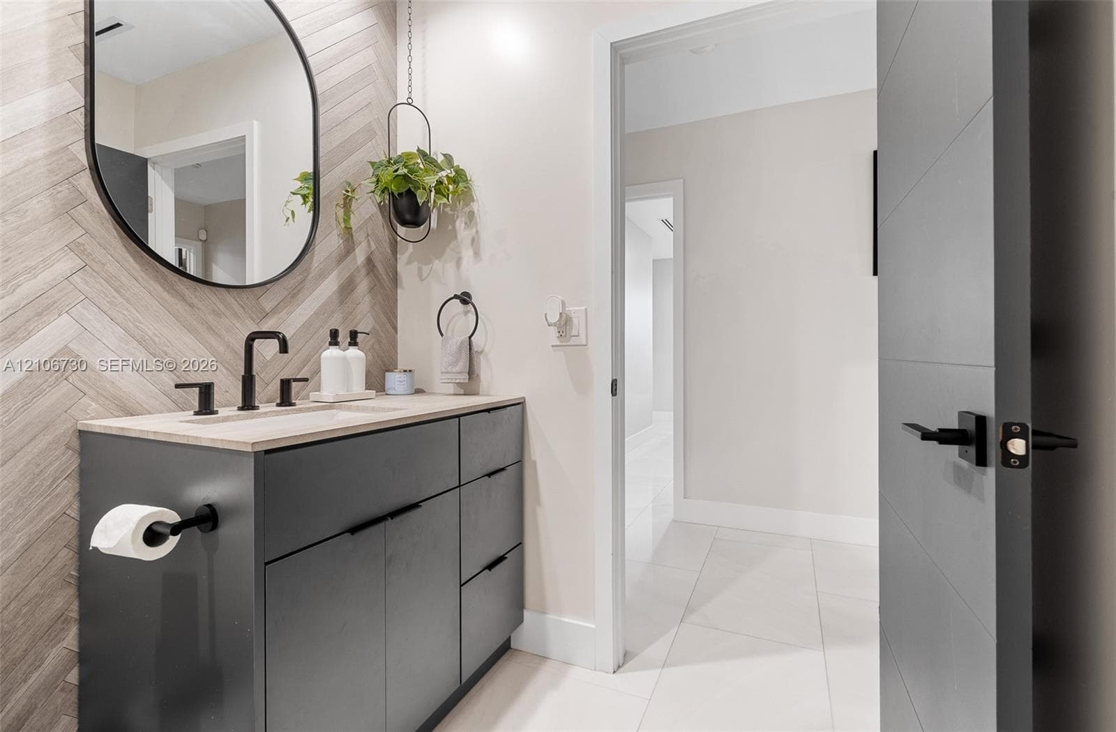Modern vanity featuring charcoal cabinetry, a light-toned stone countertop, matte black fixtures, an arched wall mirror, and a herringbone-patterned tile backsplash