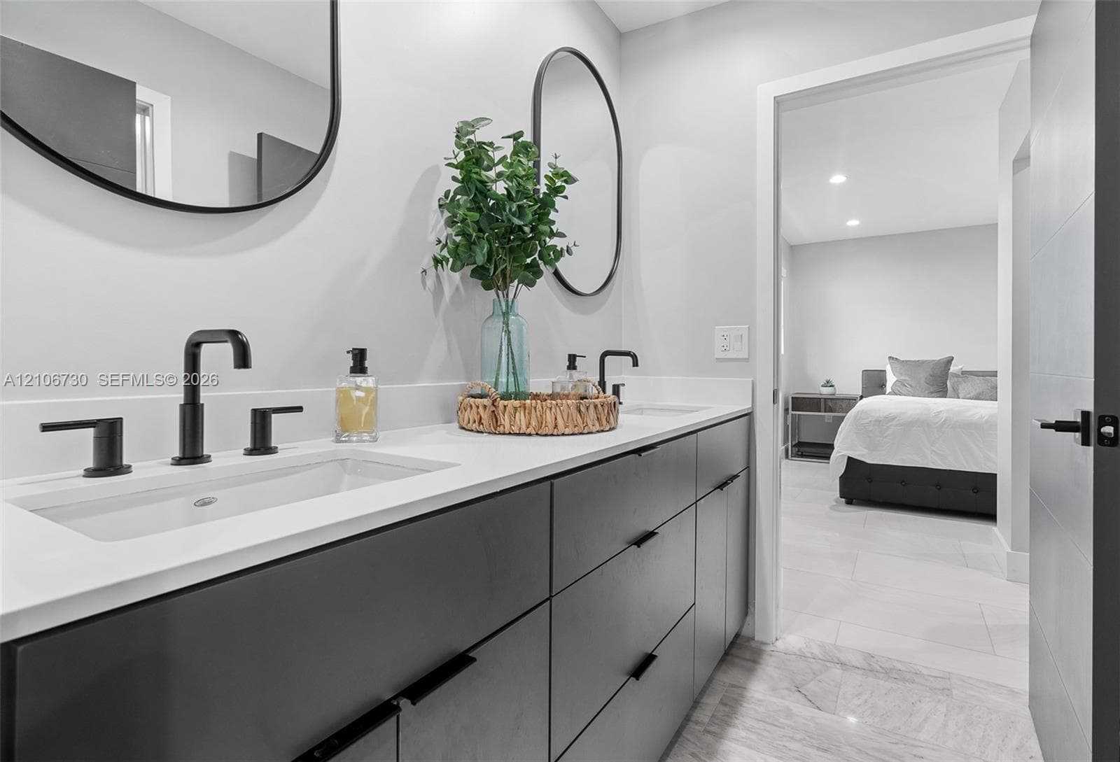 Double vanity with charcoal-toned cabinetry, light-toned stone-surface countertop, dual rectangular undermount sinks, matte black plumbing fixtures, and two oval-shaped mirrors