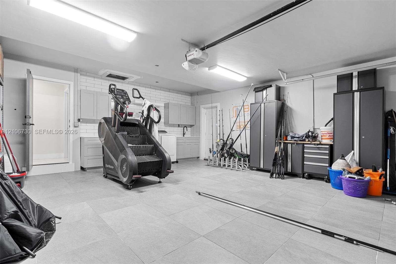 Garage featuring large-format tile flooring, built-in light grey cabinetry, a subway tile backsplash, and overhead fluorescent lighting