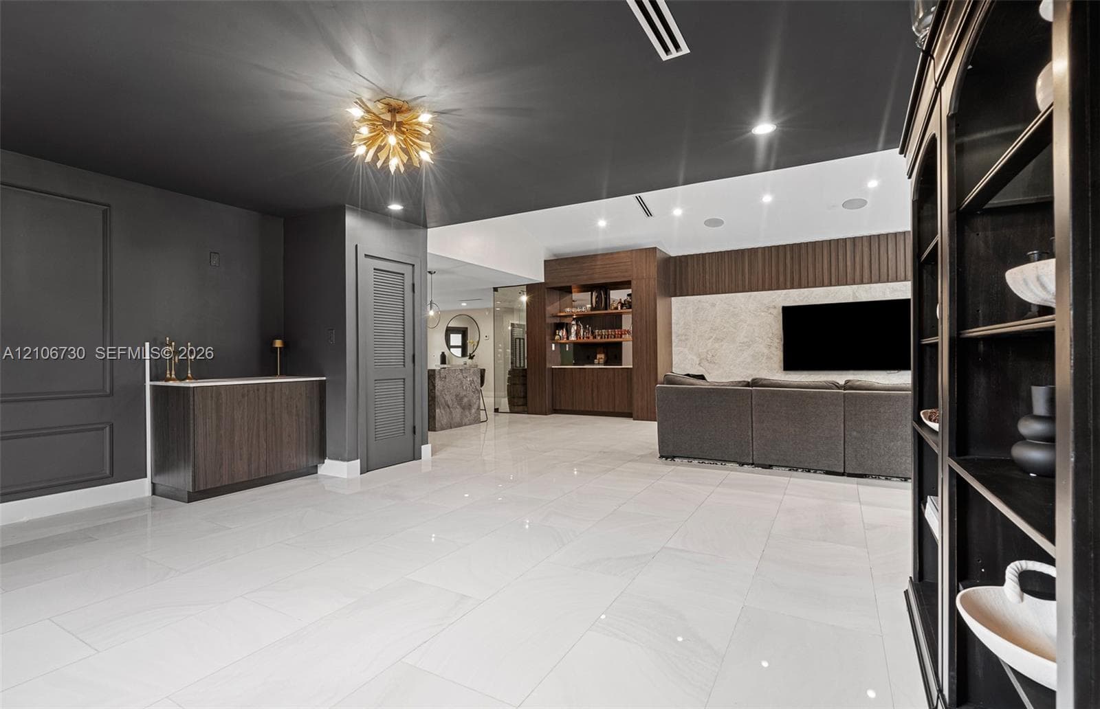 Large-format porcelain tile flooring, dark-toned wood cabinetry with integrated wet bar, textured stone accent wall, recessed lighting, and contemporary gold-tone statement chandelier