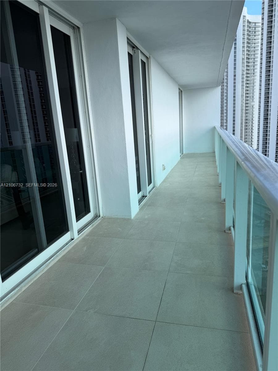 Covered balcony featuring large-format ceramic tile flooring, sliding glass door entry, and glass panel railing with a metal handrail