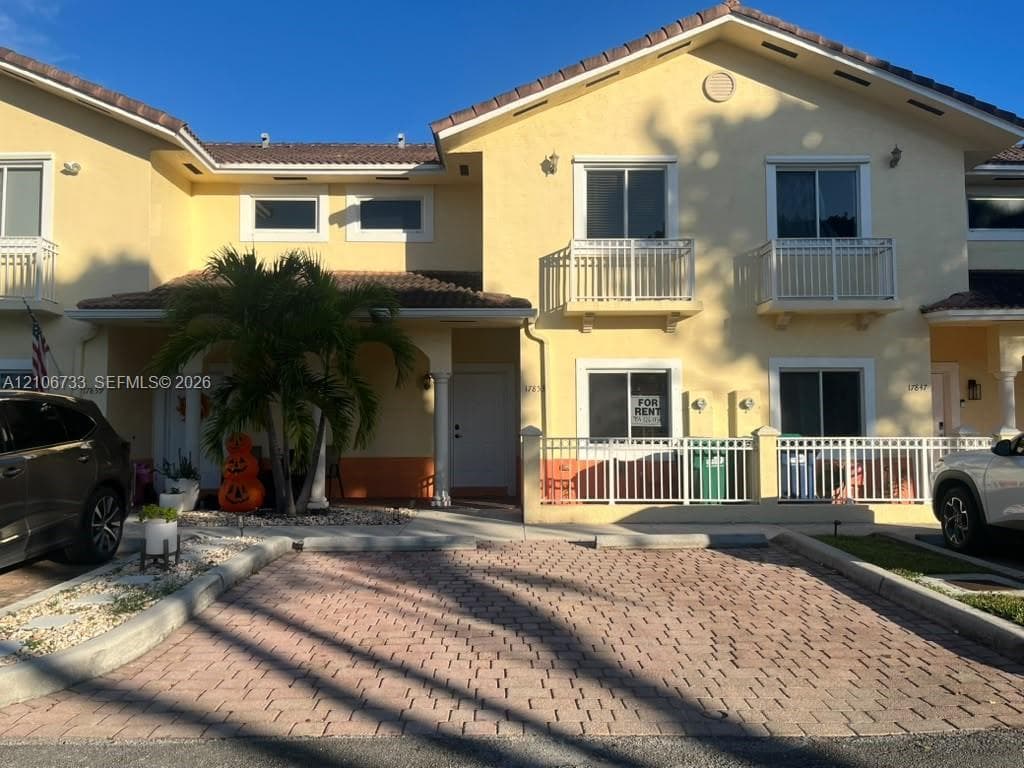 Two-story stucco townhouse exterior featuring a tiled roof, paver driveway, second-level balconies with metal railings, and a fenced front patio area
